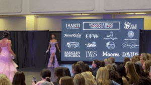 Runway models showcasing elegant gowns at the Modern Bride Wedding Show, with a large LED screen displaying sponsor logos in the background.