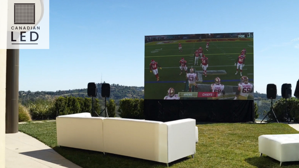Outdoor LED screen displaying a football game with white sofas and speakers set up for a backyard viewing event under a clear blue sky.