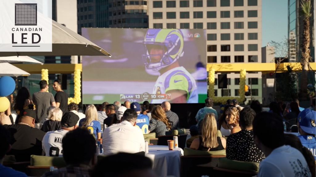 Outdoor LED screen displaying a football game to a crowd of spectators at a rooftop viewing party in an urban setting.Outdoor LED screen displaying a football game to a crowd of spectators at a rooftop viewing party in an urban setting.
