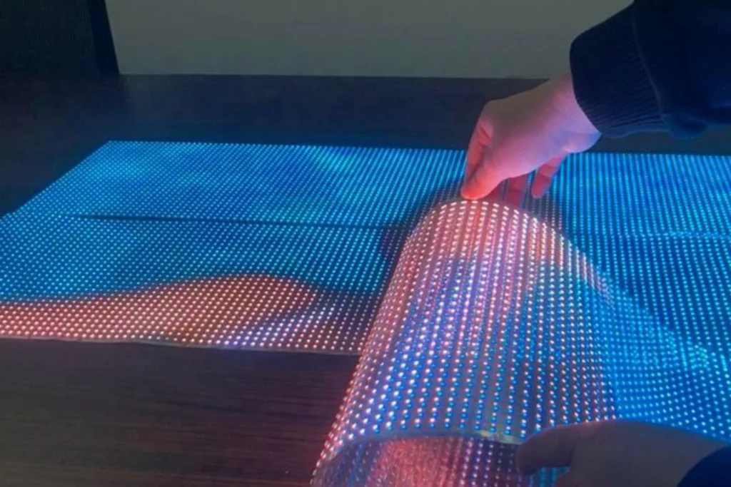 Hands holding and bending a flexible transparent LED screen panel with blue and red lights.