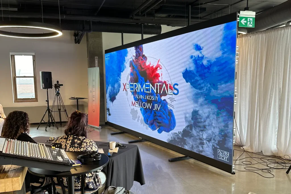 Indoor event with a large LED display showing the title "Xperimentals" and two attendees seated at a table with audio controls.